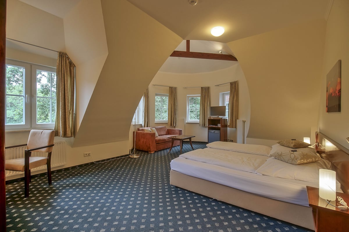 Quarto de hotel com uma cama grande, uma escrivaninha, uma cadeira e um sofá. Há janelas com cortinas, um ventilador de teto e um quadro na parede.