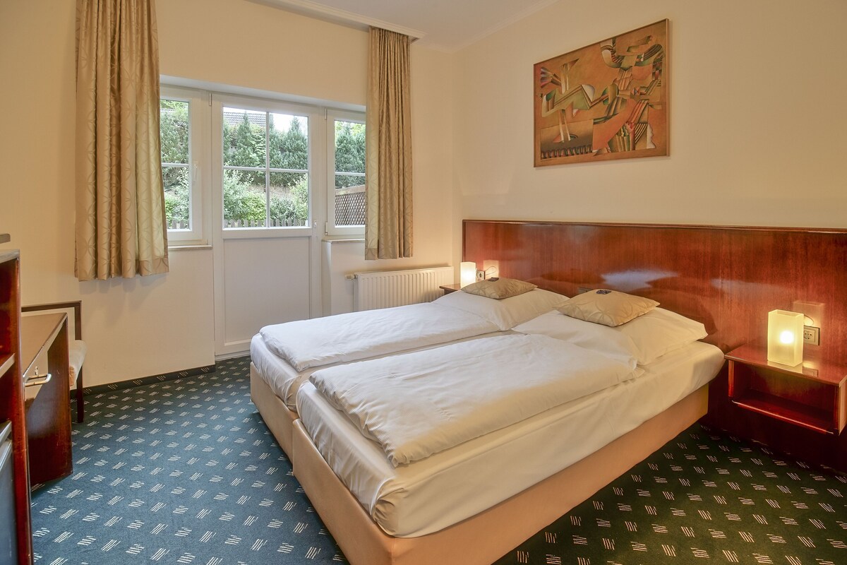 Un lit double avec du linge de lit blanc, une tête de lit en bois, une table de chevet avec une lampe, un radiateur et une fenêtre avec des rideaux.