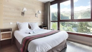 A hotel room with a large bed, wooden panel walls, two bedside lamps, a view of a valley, and a window with curtains. - Wifi, 100m from resort center, swimming pool, balcony, tv, ski locker, 58m², Flaine (Flaine)
