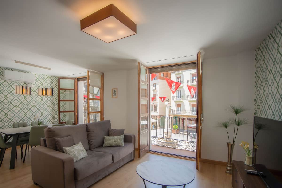Un salon moderne avec un canapé, une table à manger et des chaises. Il y a un balcon décoré de drapeaux rouges, d’où l’on peut admirer la vue sur les immeubles.