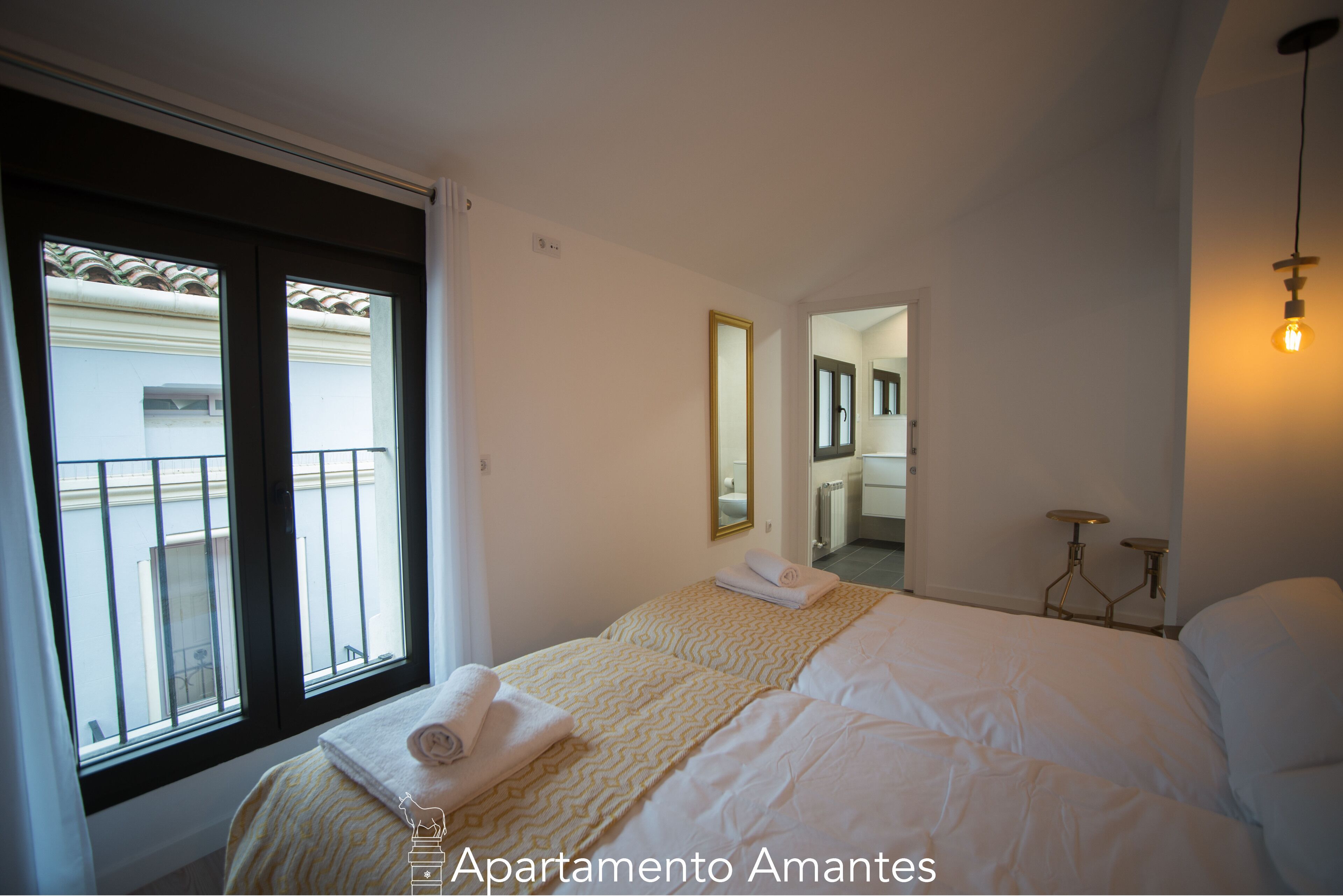 A bedroom with a bed, a window, a mirror, and a bathroom visible through an open door.