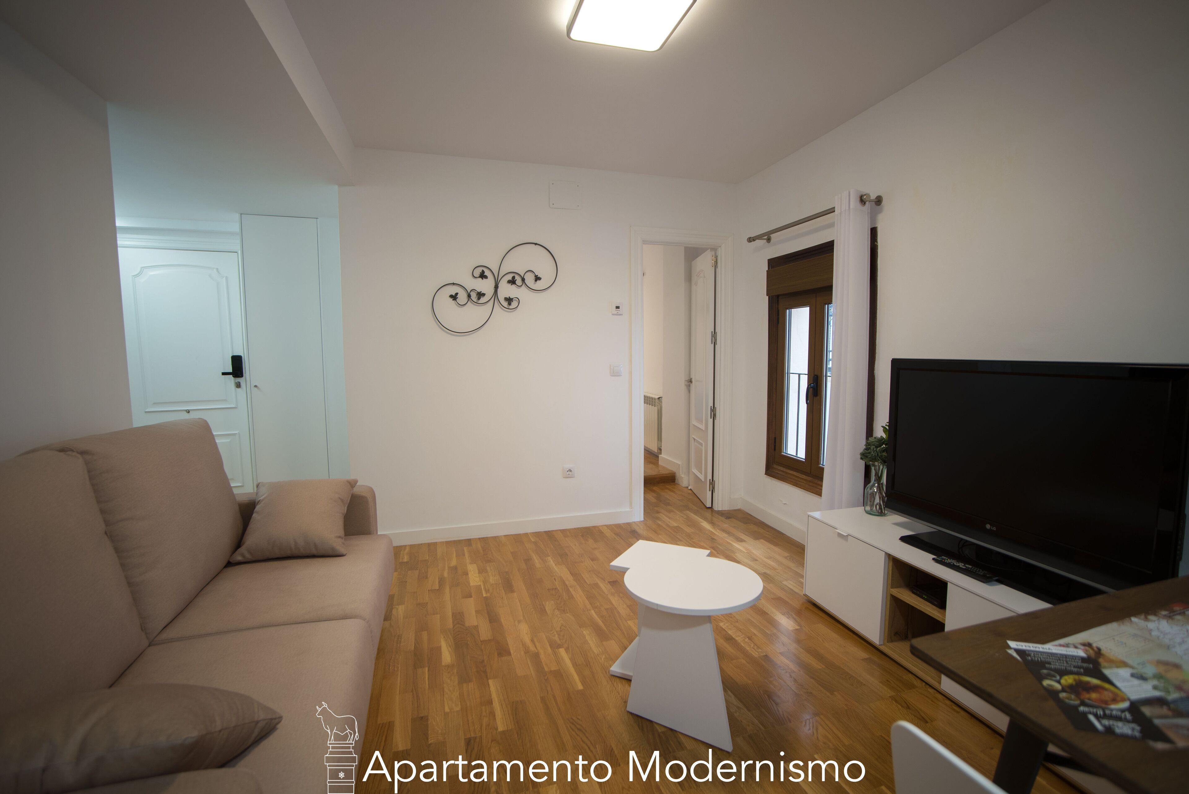 A modern apartment with a beige sofa, a small white table, a flat-screen TV, and a wooden floor.