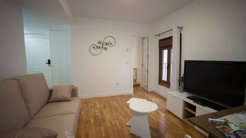 A modern apartment with a beige sofa, a small white table, a flat-screen TV, and a wooden floor.