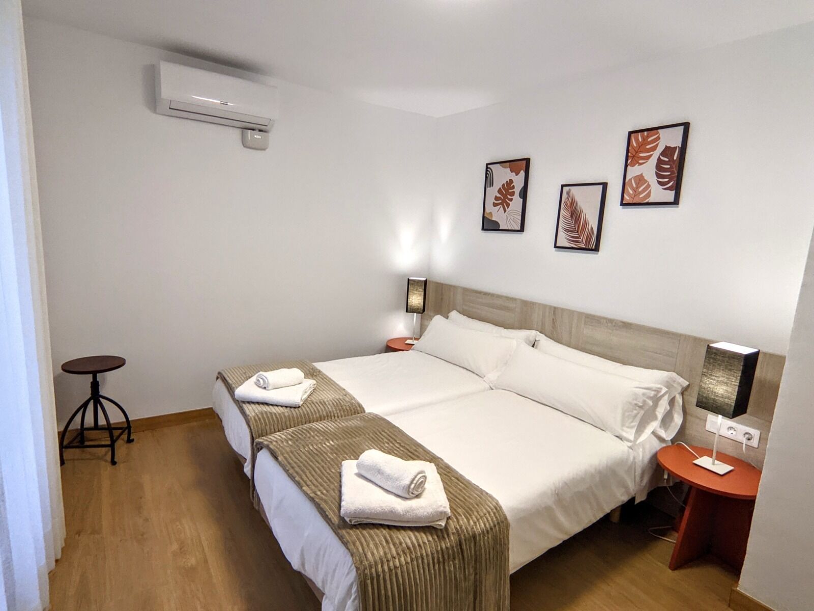 A hotel room with two beds, a small table, a lamp, and framed artwork on the wall.
