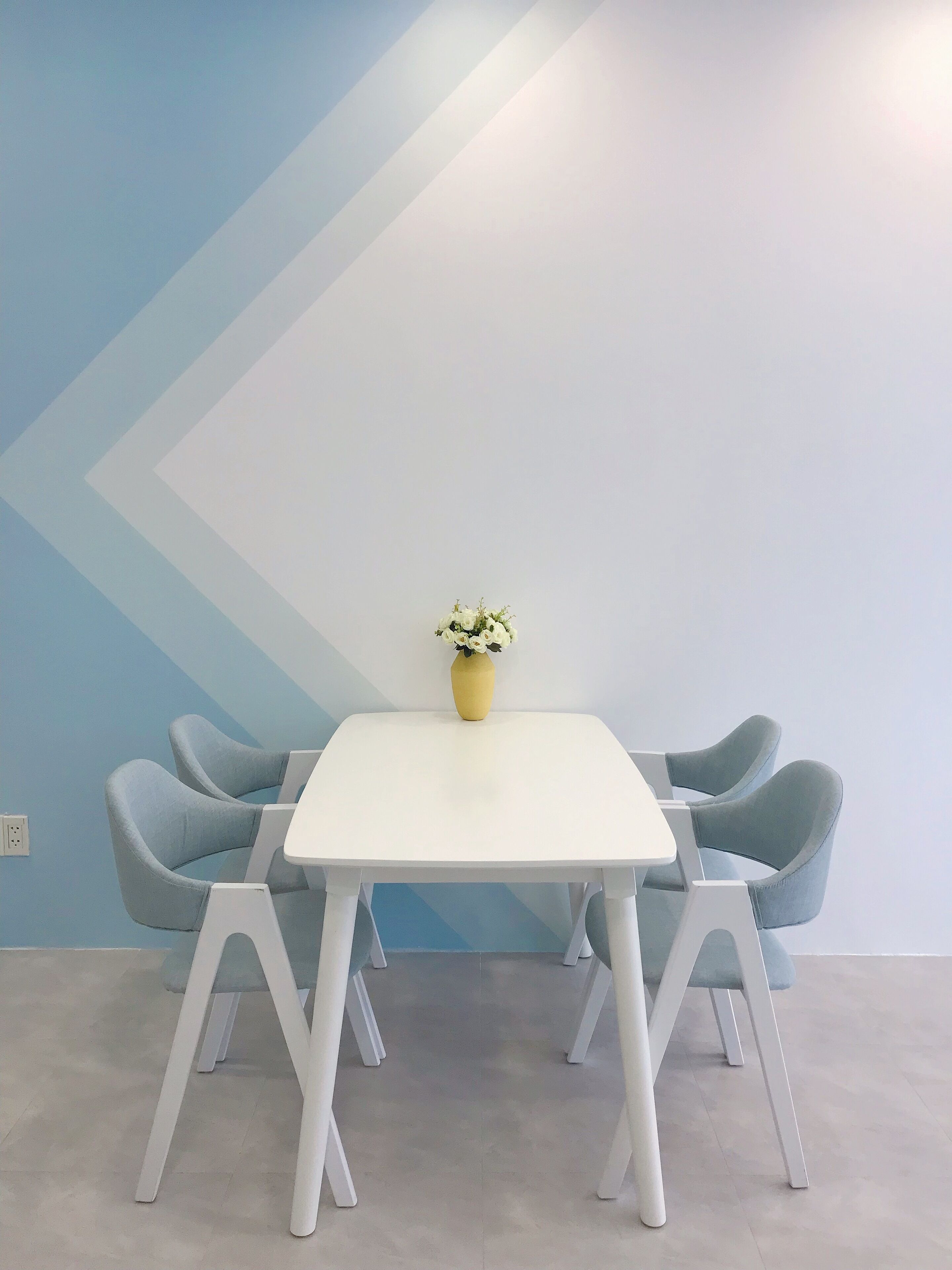 Une salle de réunion moderne avec une table blanche et des chaises bleues, un vase jaune orné de fleurs et un mur à motifs géométriques.