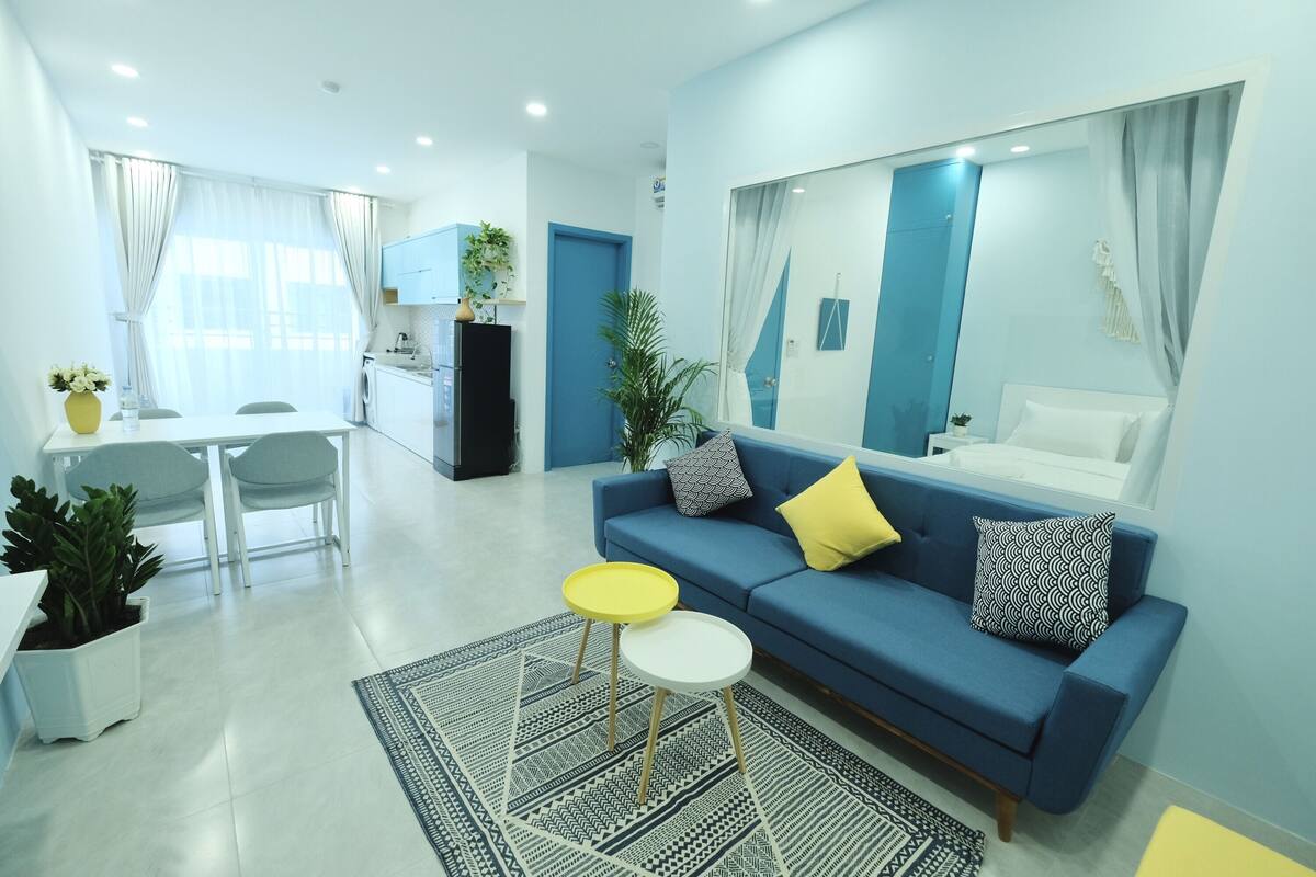 Ein modernes Wohnzimmer mit einem blauen Sofa, gelb-weißen Beistelltischen, einem großen Spiegel und einem Essbereich mit einem weißen Tisch und Stühlen.