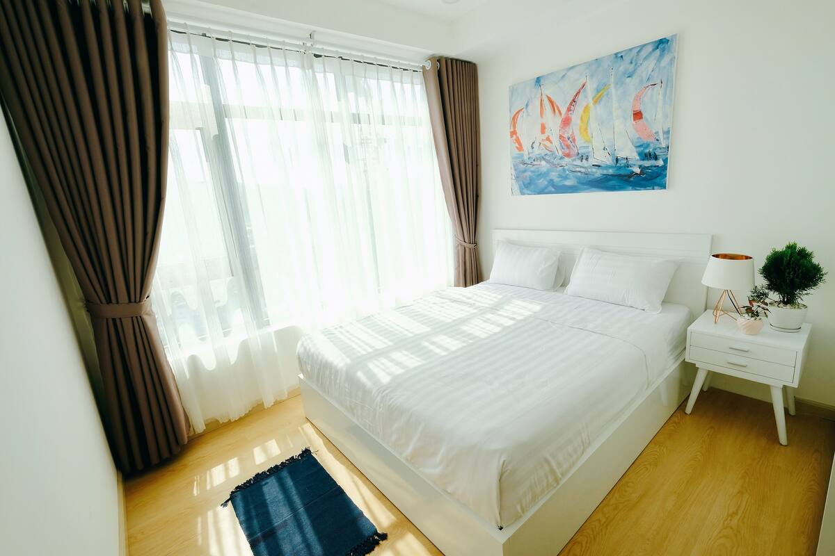 Ein Schlafzimmer mit Bett, Vorhängen, einem Gemälde, einem Nachttisch und einer Pflanze.