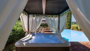 A hot tub under a covered area with a view of a swimming pool and greenery.