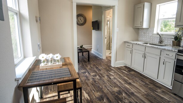 Una cocina con suelo de madera, armarios blancos, un lavabo y un comedor con mesa y sillas.