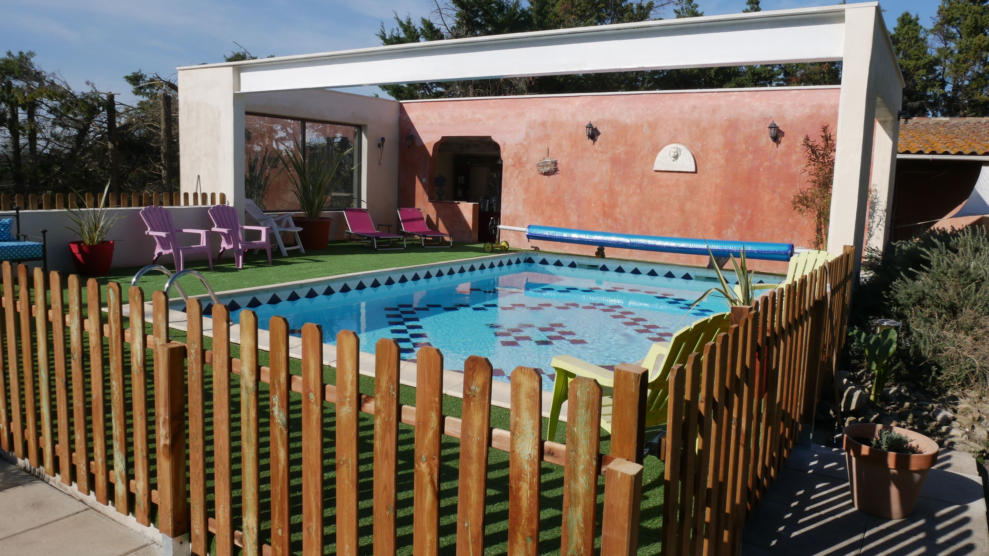 A swimming pool surrounded by a wooden fence, with a patio area featuring chairs and a potted plant.