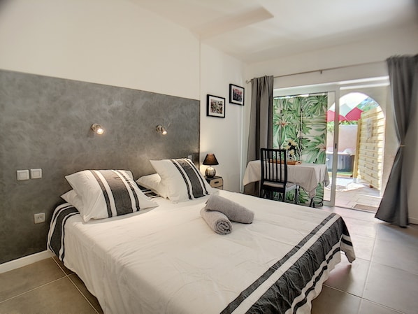 Une chambre à coucher comprenant un lit, une table de chevet, une lampe et donnant sur une terrasse extérieure avec une table et des chaises.