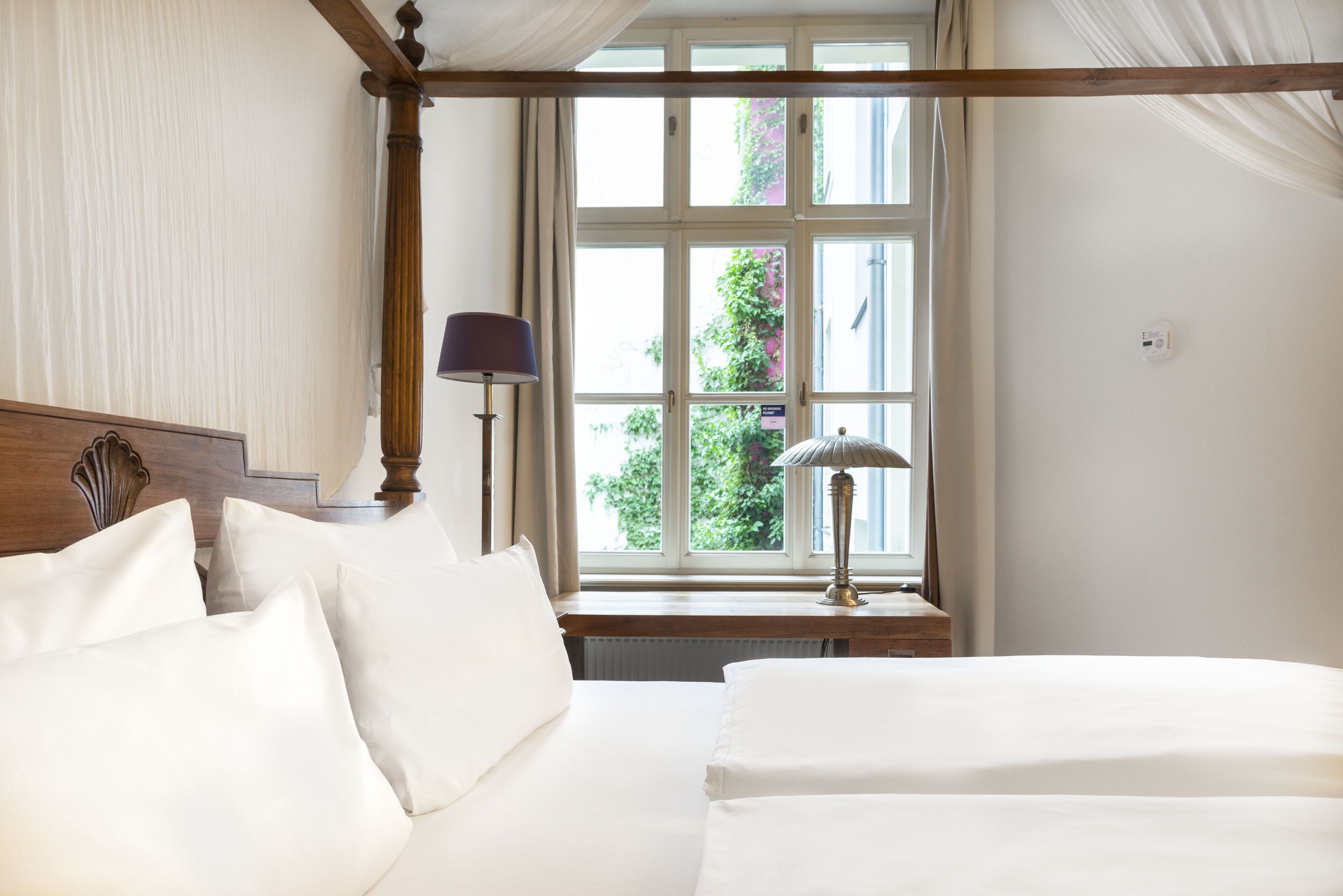 A four-poster bed with white linens, a bedside lamp, and a window with a view of greenery.