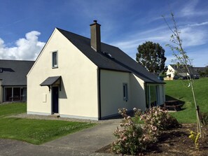 A small, single-story house with a sloped roof and a chimney, surrounded by a well-maintained lawn and a few flowering plants. - Seamount Holiday Village No 24 (Courtown)