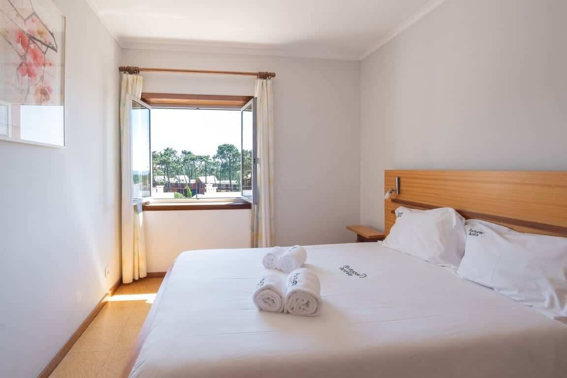 Una habitación de hotel con una cama, cabecera de madera, mesita de noche, ventana con cortinas y vistas a árboles y edificios.