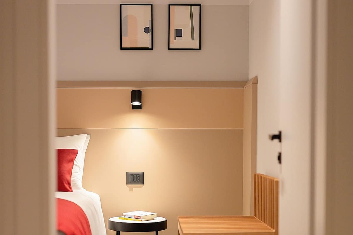 Ein Hotelzimmer mit Bett, Nachttisch und einem kleinen Holzsessel. Über dem Bett hängen zwei gerahmte Bilder an der Wand.
