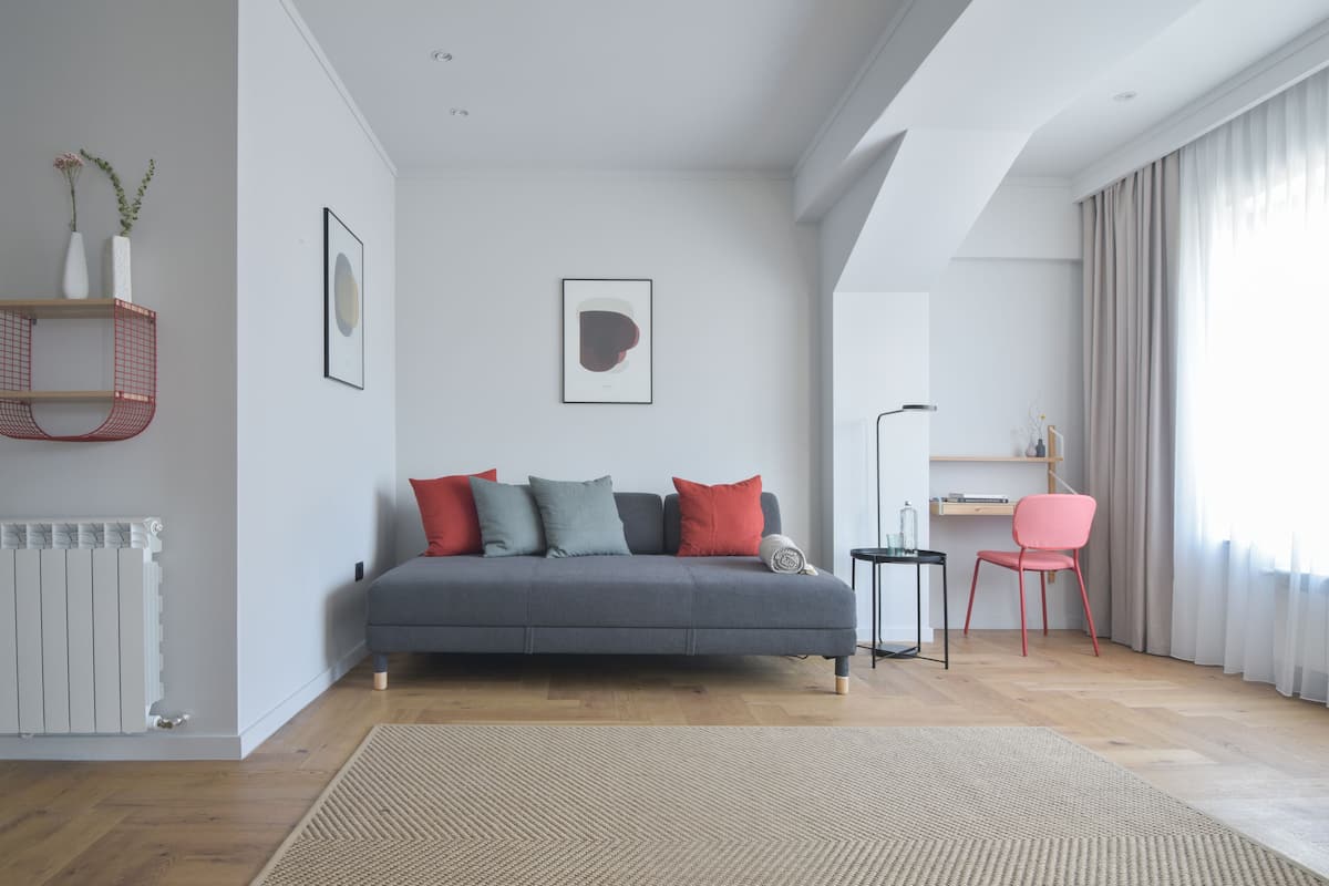 Ein modernes Wohnzimmer mit einem grauen Sofa, roten und grauen Kissen, einem Holzboden, einem roten Sessel, einer schwarzen Stehlampe und einem gerahmten Bild an der Wand.