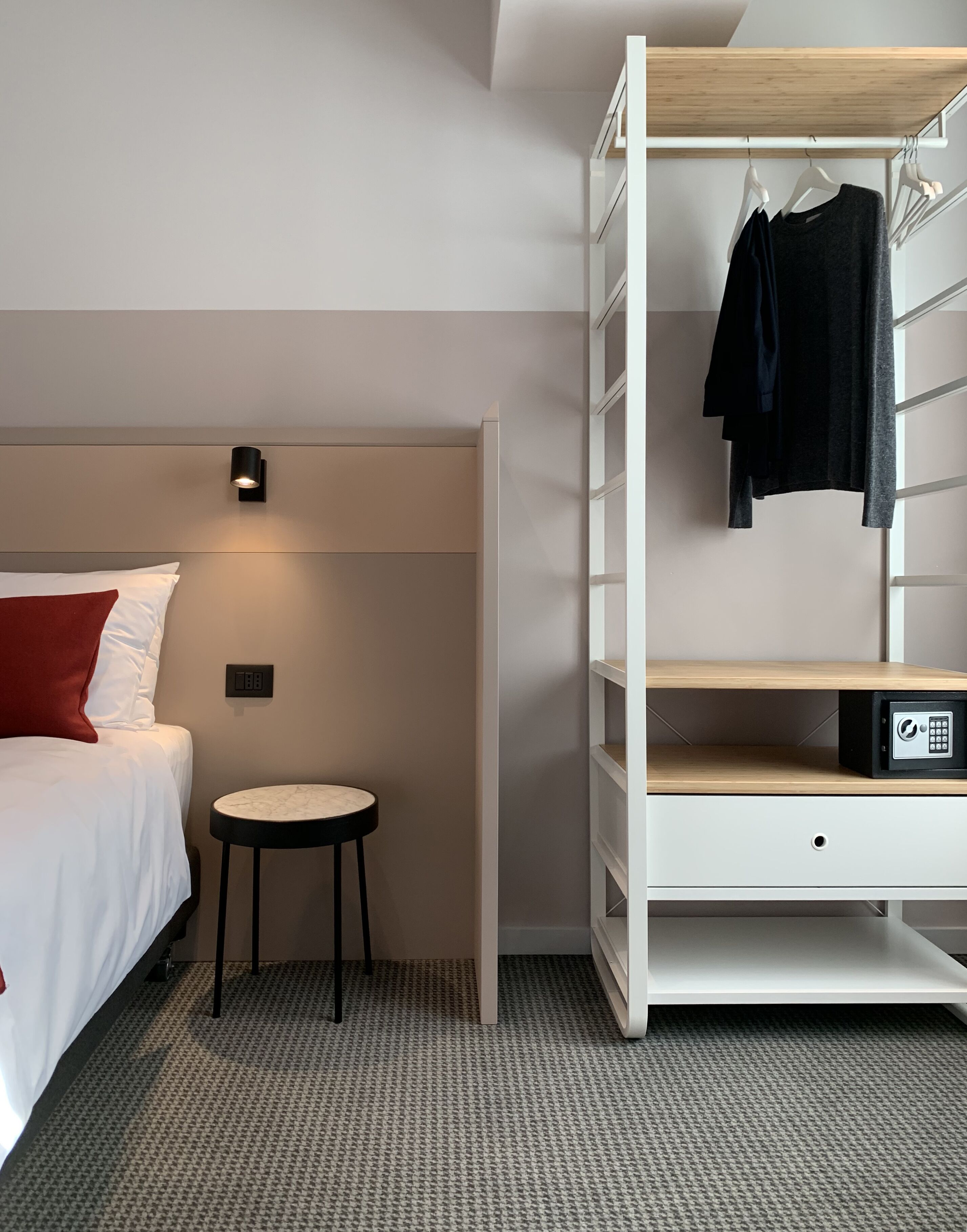 A hotel room with a bed, a small round table, a wall-mounted light, a wardrobe with hanging clothes, and a safe.