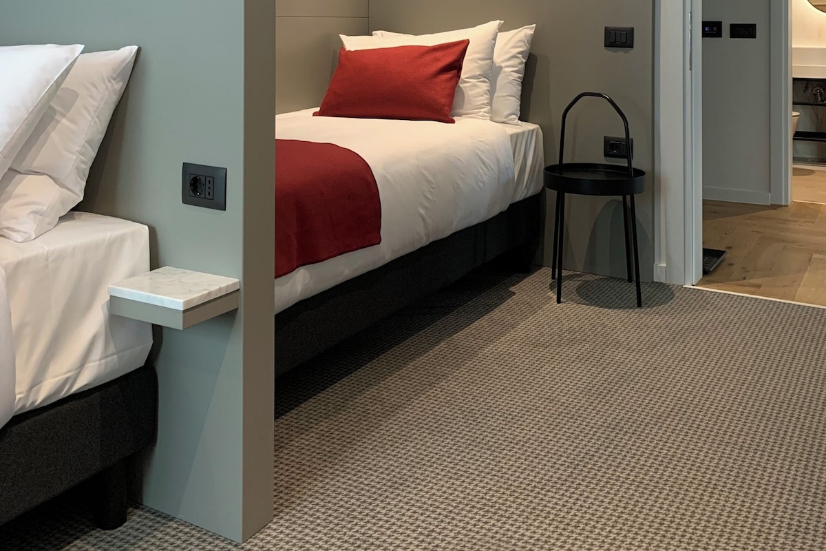 A modern hotel room with a bed, a small table, and a chair. There is a partition wall and a door leading to another room.