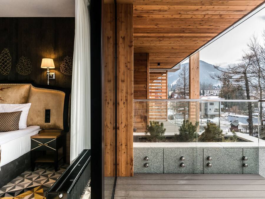 Une chambre d’hôtel moderne équipée d’un lit, d’une table de chevet, d’une lampe et d’un balcon offrant une vue sur la ville et les montagnes.