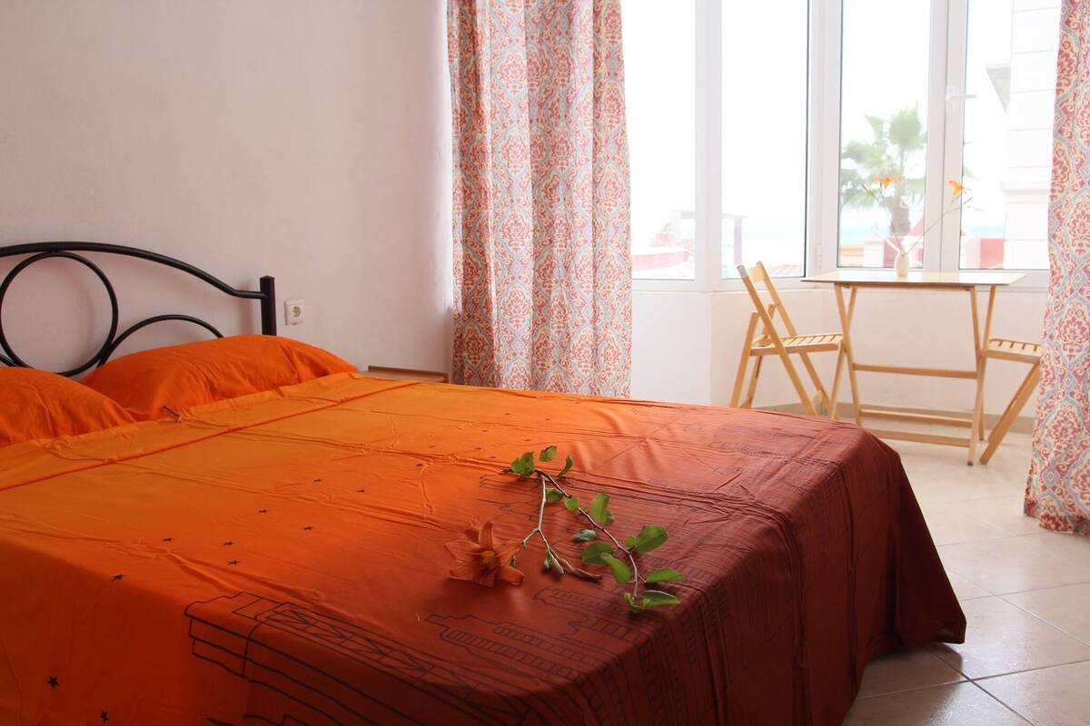 Una cama con una colcha naranja, una mesita con una silla y una ventana con cortinas.