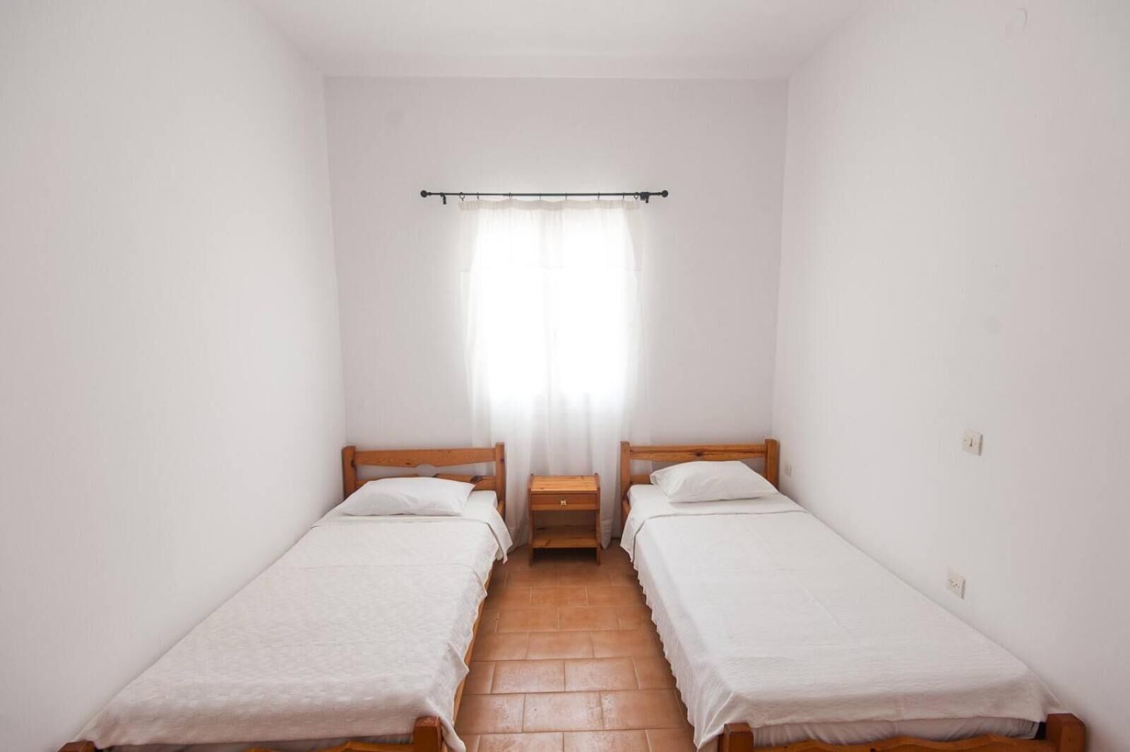 Una habitación pequeña y limpia con dos camas individuales, un cabecero de madera, una mesita de noche y una ventana con cortinas traslúcidas.
