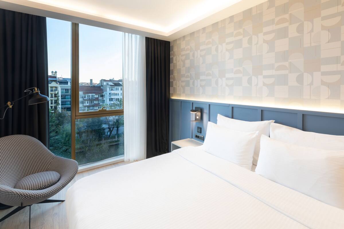 Een moderne hotelkamer met een groot bed, een stoel, een nachtkastje en uitzicht op de stad door een groot raam.