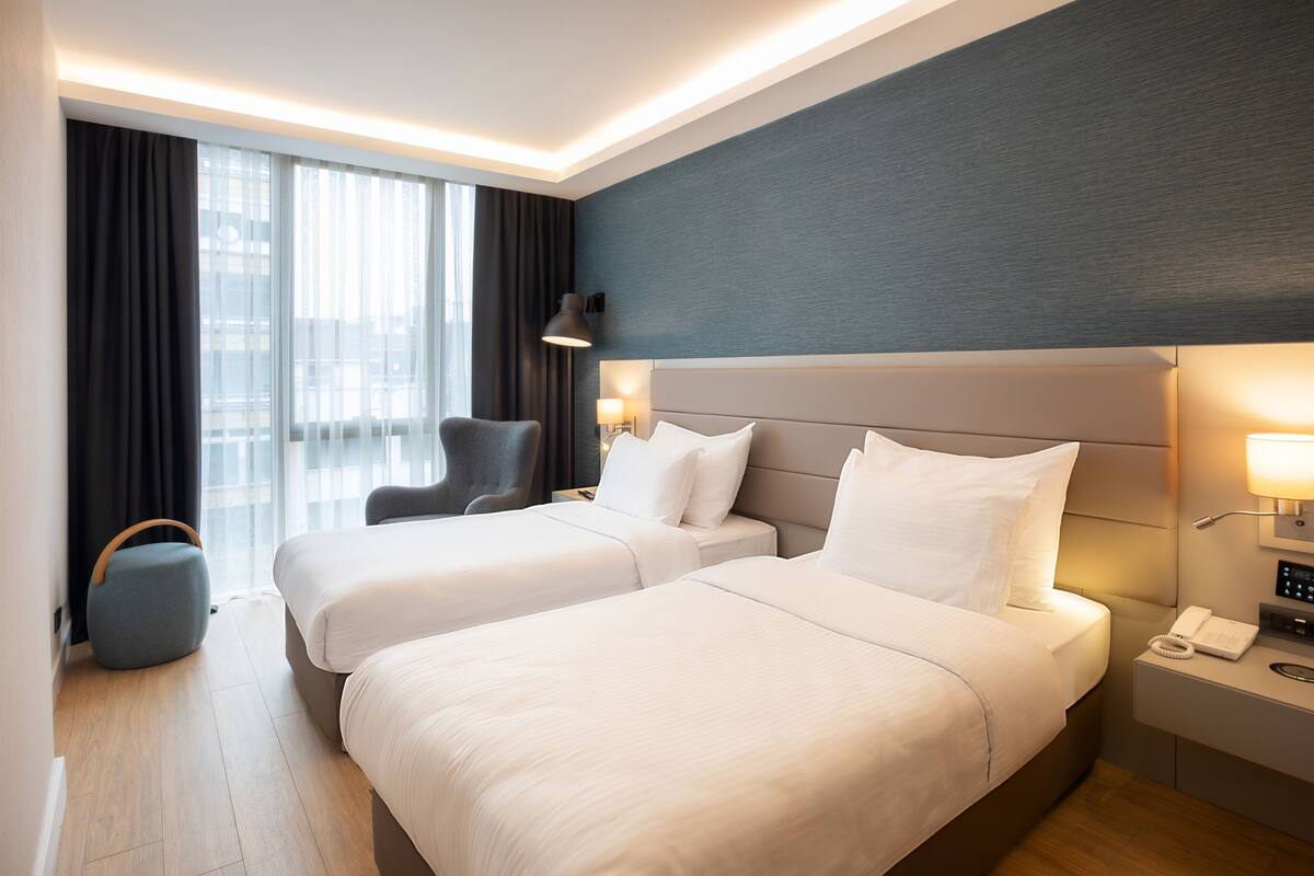 Een hotelkamer met twee bedden, een bureau, een stoel, een lamp en uitzicht op de stad door grote ramen.
