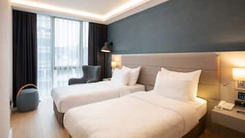 A hotel room with two beds, a desk, a chair, a lamp, and a view of the city through large windows.