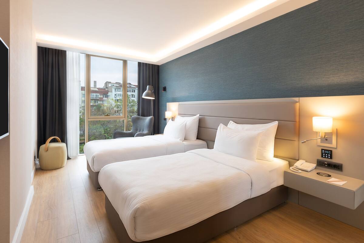 Een hotelkamer met twee bedden, een bureau met een lamp, een stoel en uitzicht op gebouwen.