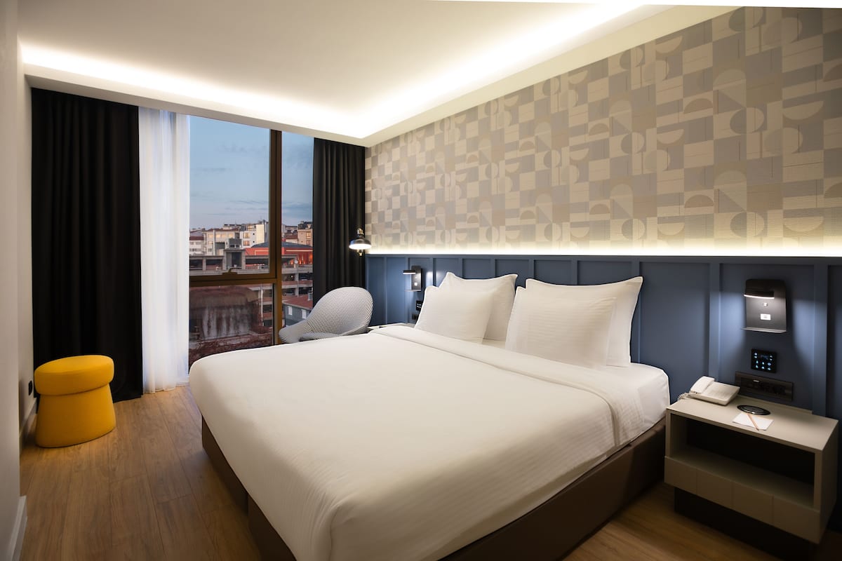 Een moderne hotelkamer met een groot bed, een nachtkastje met telefoon en lamp, een klein geel poefje en uitzicht op de stad door grote ramen.