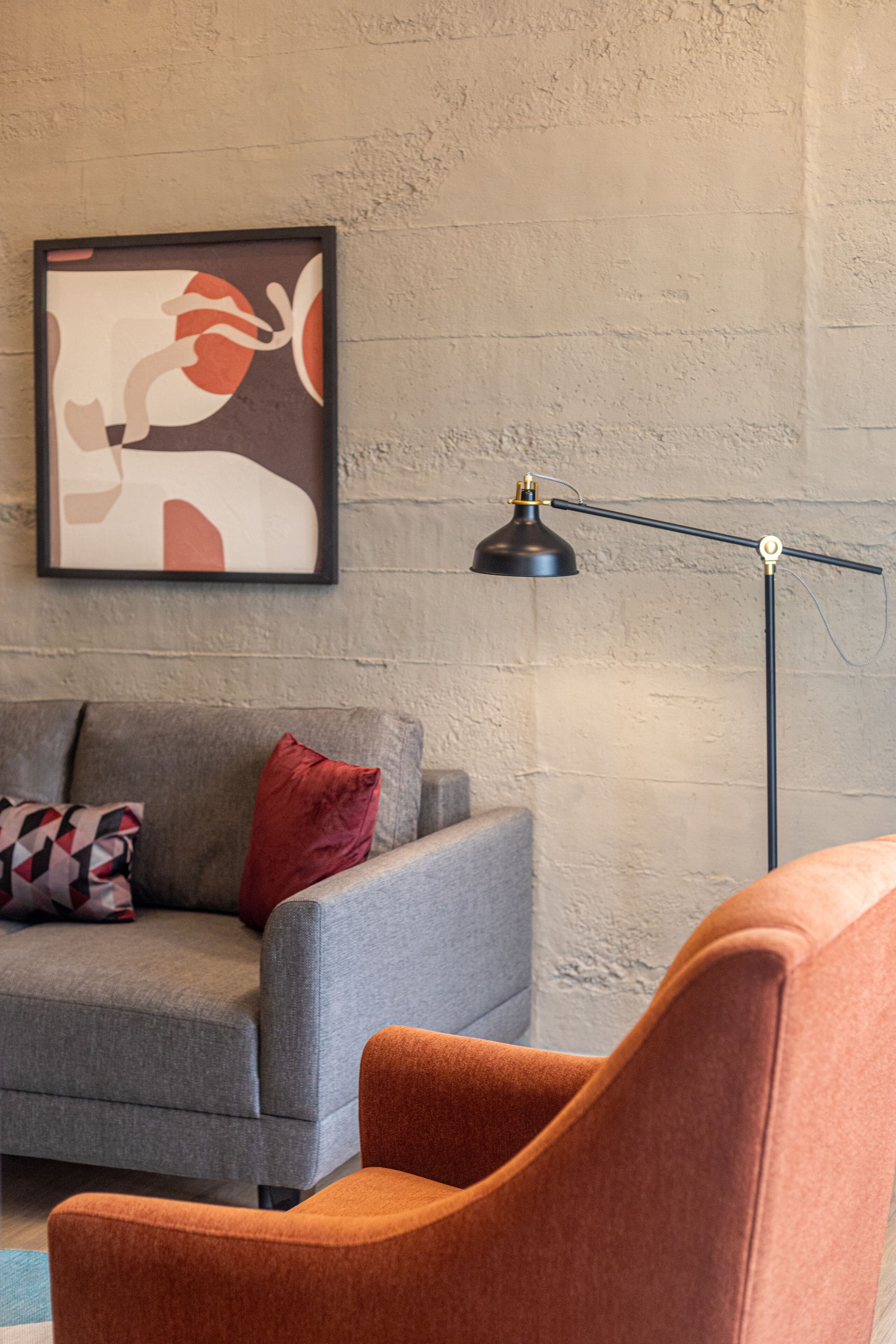 A living room with a grey sofa, a red pillow, an orange armchair, a floor lamp, and a framed abstract artwork on the wall.