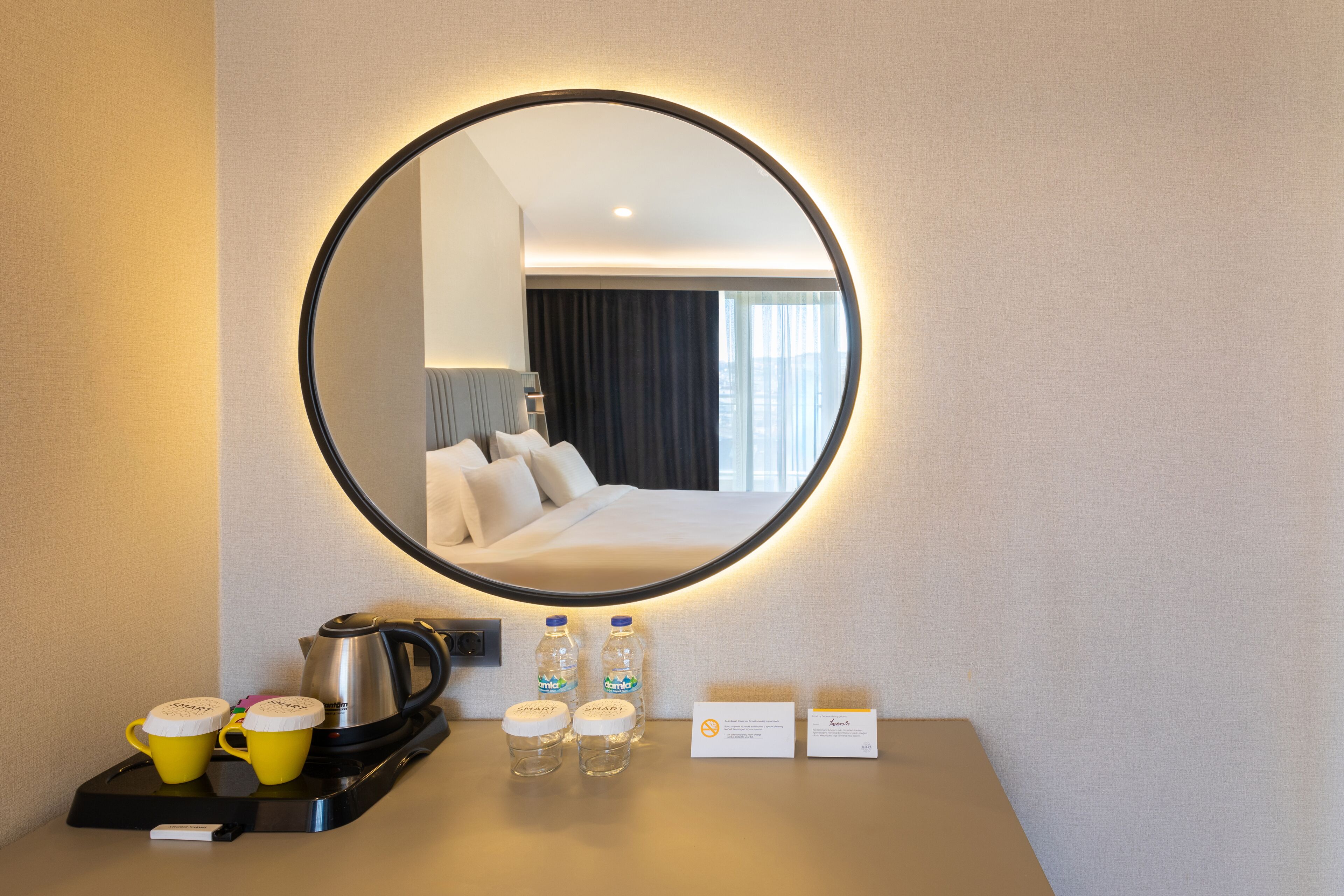 A hotel room with a round mirror, a bed, a coffee maker, two yellow mugs, two water bottles, and a tray with a kettle and a sign.
