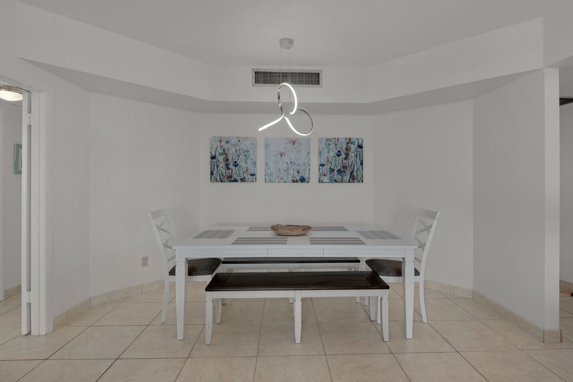 Un comedor con una mesa y sillas blancas, una lámpara colgante y tres cuadros con motivos florales en la pared.