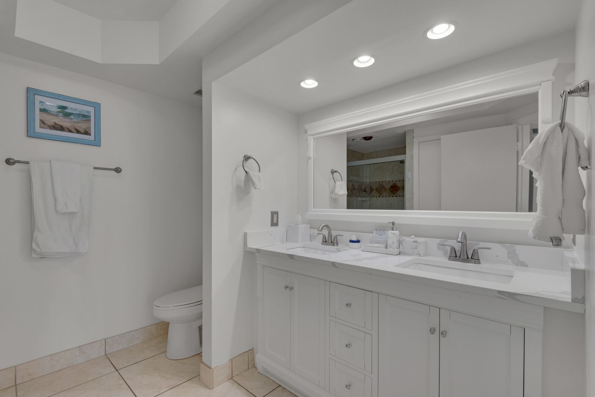 A modern bathroom with a white sink, vanity, and toilet. There is a towel rack with towels, a framed picture on the wall, and recessed ceiling lights.