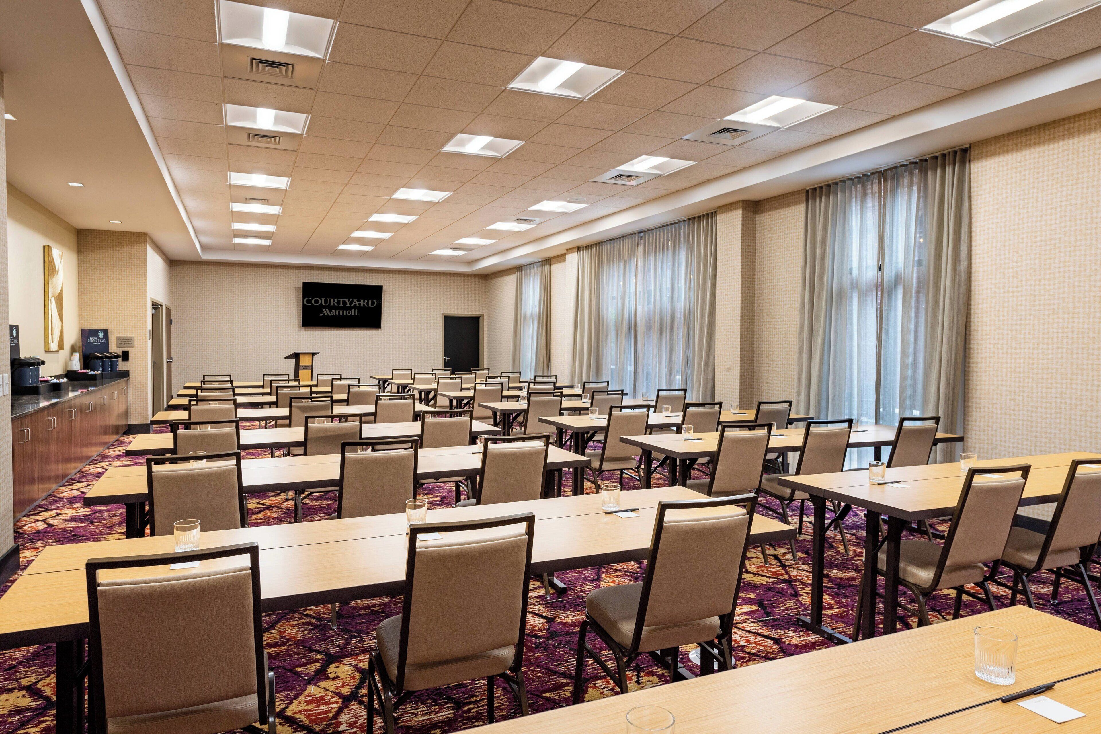 Un salón de conferencias con varias mesas y sillas, piso alfombrado y un amplio ventanal con cortinas.