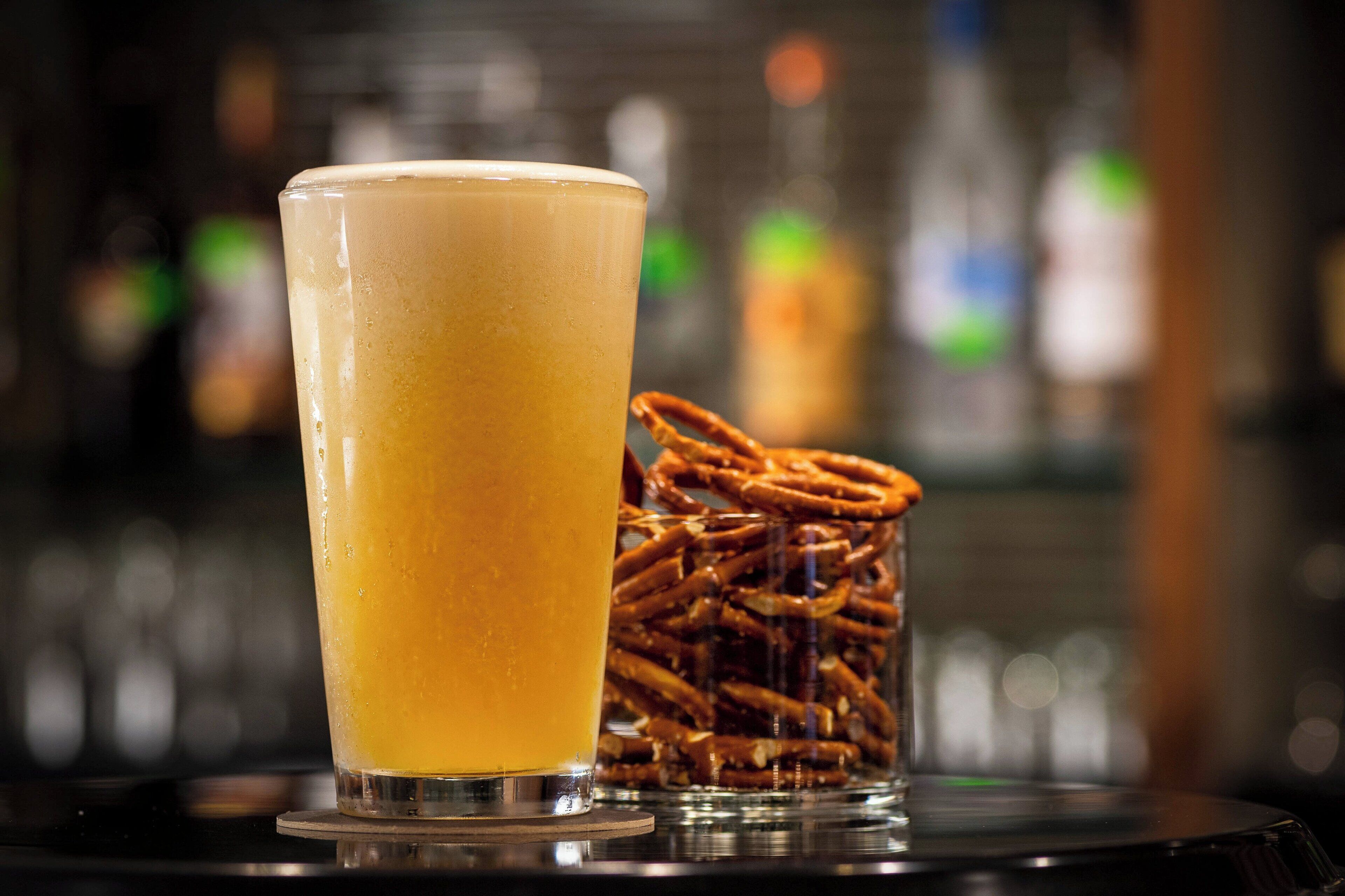A tall glass of orange beverage on a bar counter with a pile of pretzels in the background.