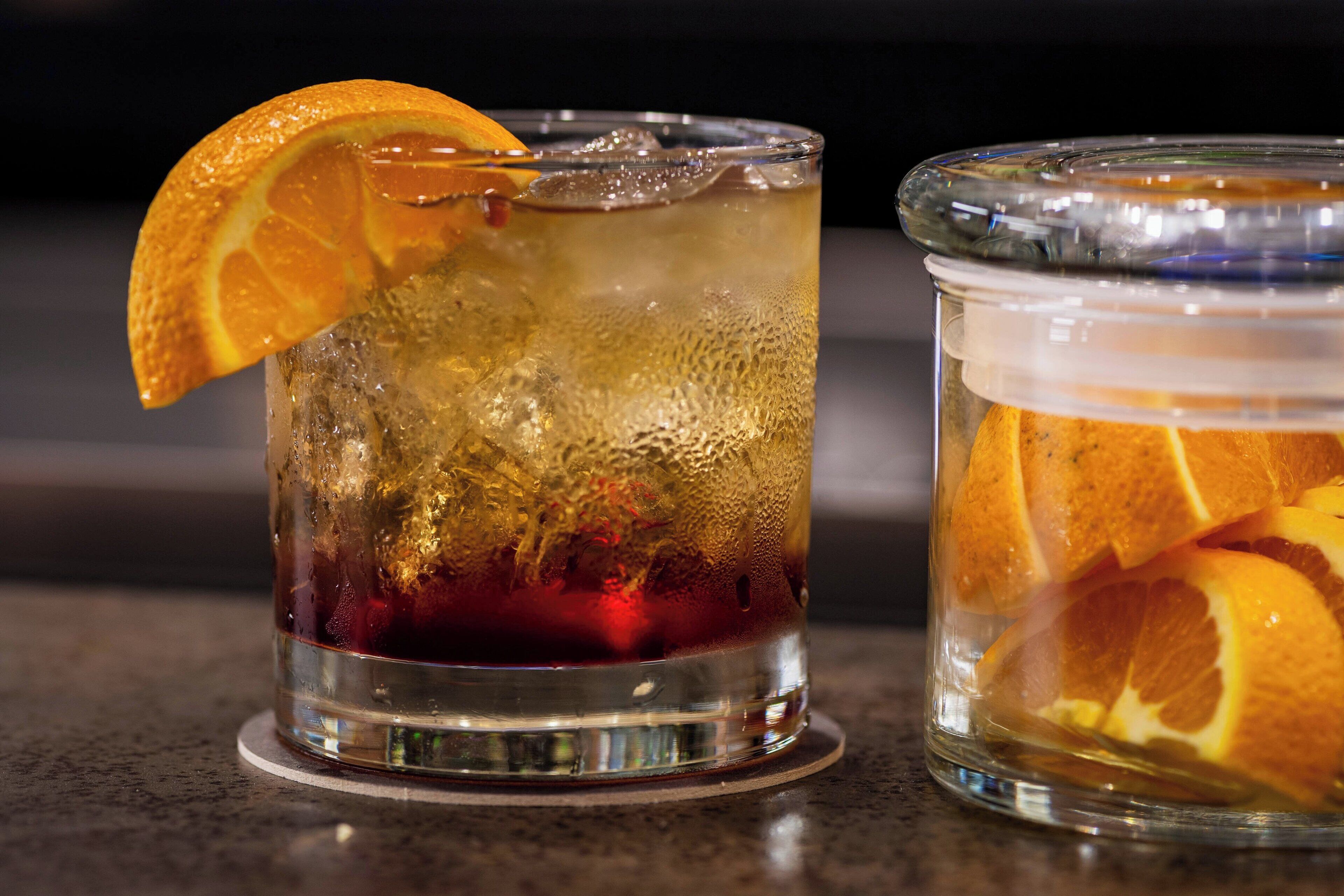 Two glasses of iced beverage with orange slices, one with a clear liquid and the other with a red liquid, on a countertop.