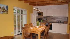 A dining area with a wooden table and chairs, a kitchen with white cabinets and appliances, and a sliding glass door leading to an outdoor area. - Haus Lutki (Burg (Spreewald))