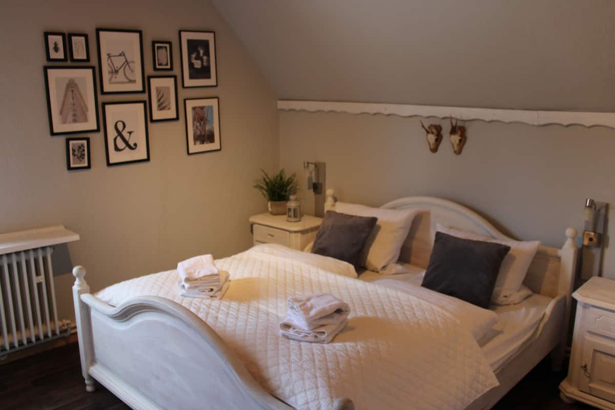 A neatly made bed with a quilted blanket, a bedside table with a lamp, and a wall decorated with framed pictures and a small plant.