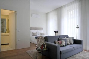 A modern living room with a grey sofa, a small round table with a vase and a lamp, and a bed with white linens in the background. - Mayor's Apartments (Porto)