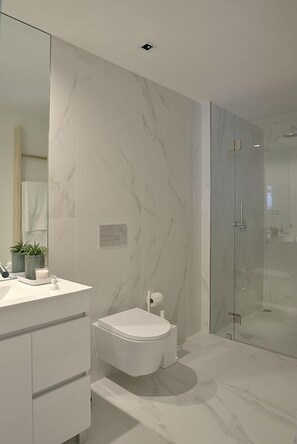 A modern bathroom with a marble floor, a white toilet, a glass shower enclosure, and a vanity with a mirror and plants. - Mayor's Apartments (Porto)