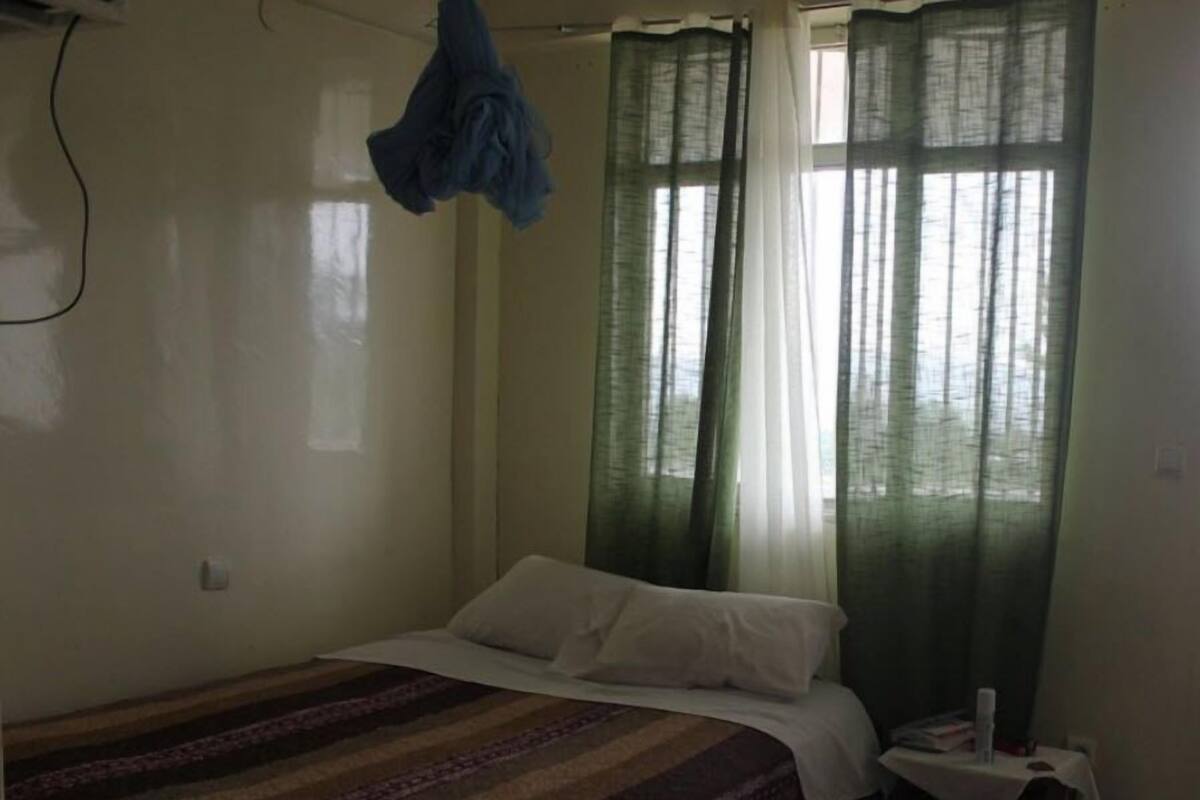 Una cama individual con una colcha a rayas, una mesita de noche con una lámpara y cortinas translúcidas en la ventana.