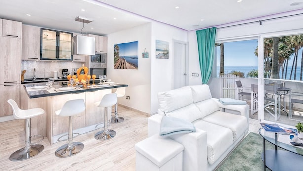 Una cocina moderna con una isla central, taburetes blancos tipo bar y una zona de comedor con sofá y mesa blancos.