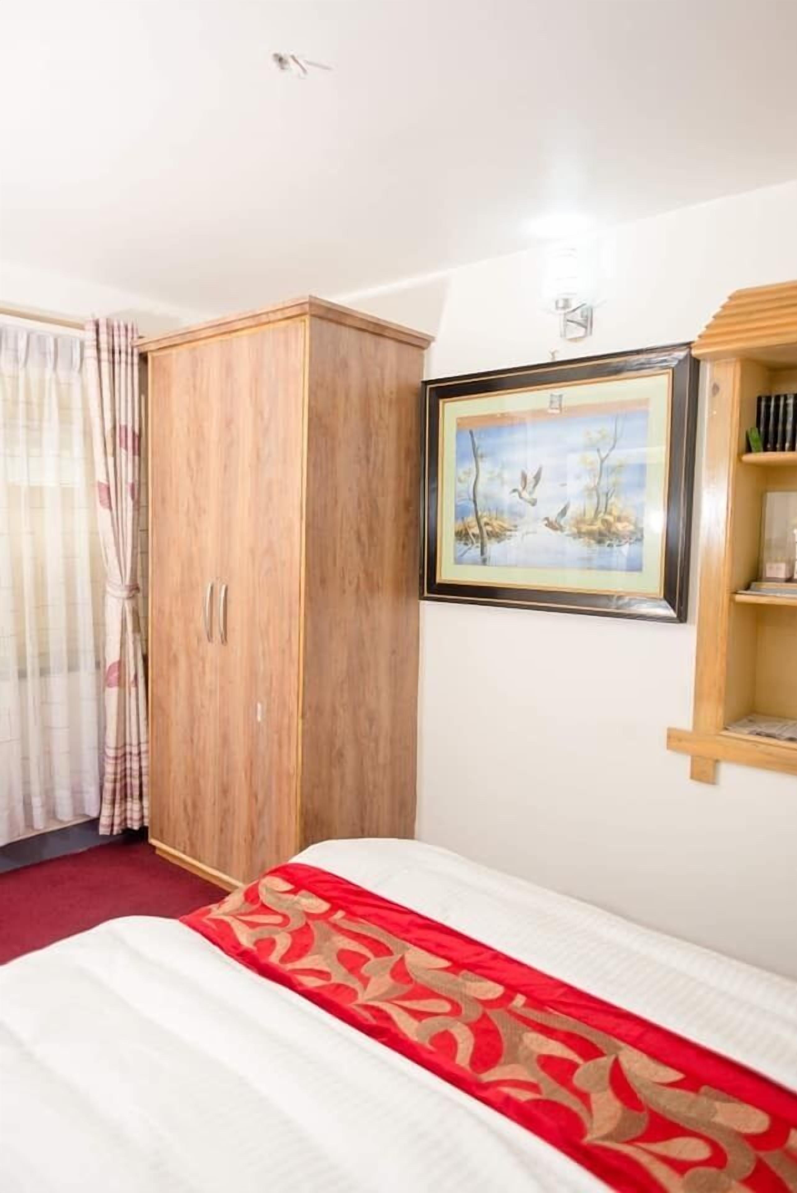 A hotel room with a bed, a wooden wardrobe, a framed picture on the wall, and a shelf with books.