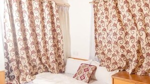 Une chambre avec un plafond en bois, des rideaux à motifs, un lit recouvert d’une courtepointe rouge et blanche, et une table de chevet en bois.
