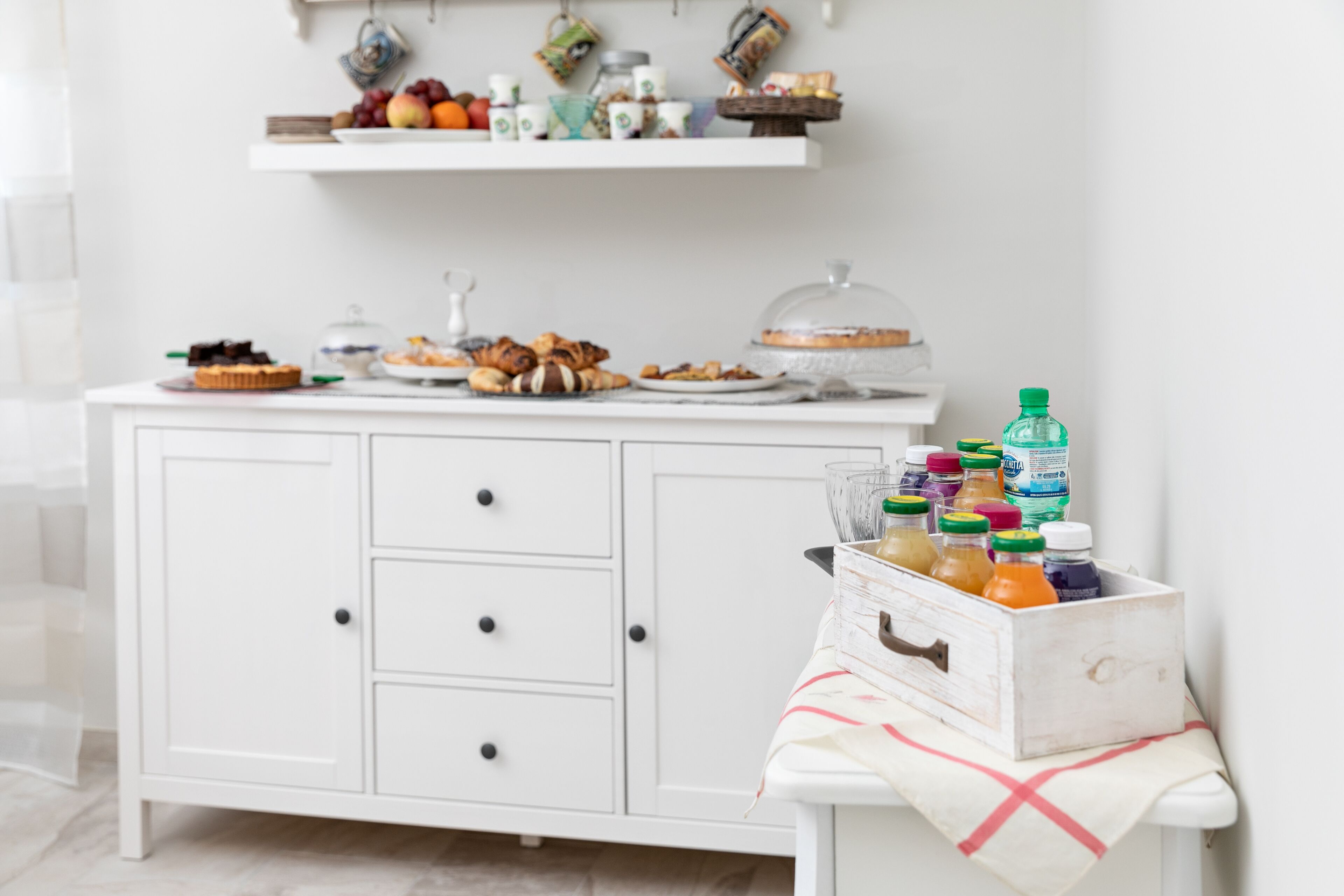 A white cabinet with drawers, a table with a cloth, and a shelf above holding various food items and drinks.