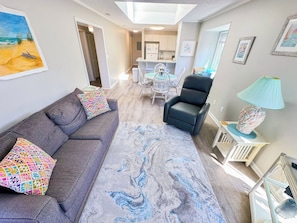 A living room with a grey sofa, colorful pillows, a patterned rug, a black armchair, a small table with a lamp, and a dining area in the background. - Tilghman Beach and Racquet Club 320 (North Myrtle Beach)