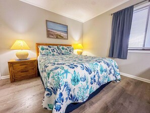 A bedroom with a bed, two bedside lamps, a nightstand, a painting on the wall, and a window with curtains. - Tilghman Beach and Racquet Club 320 (North Myrtle Beach)