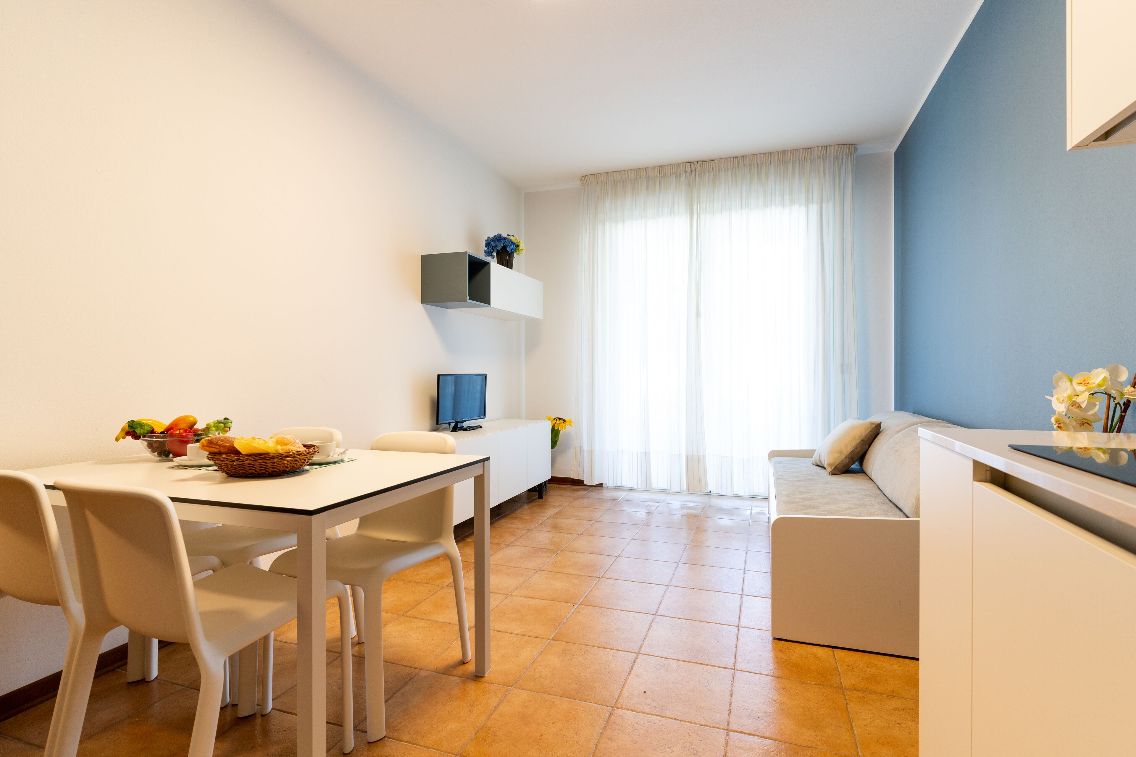 A modern living room with a sofa, a dining table with chairs, a TV, and a kitchen area with a counter and cabinets.