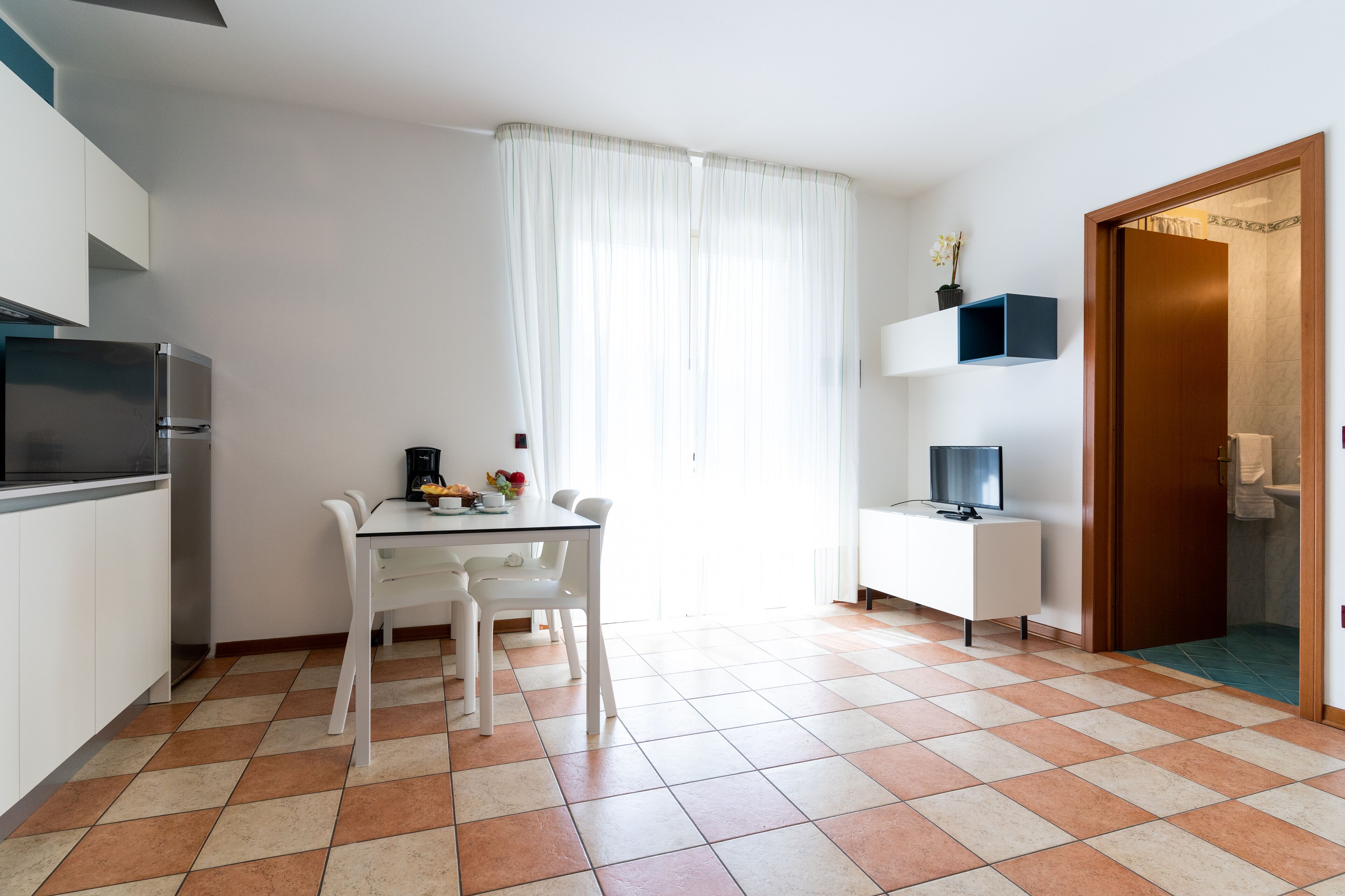 A modern kitchen with white cabinets, a black refrigerator, a dining table with chairs, and a TV on a white stand.